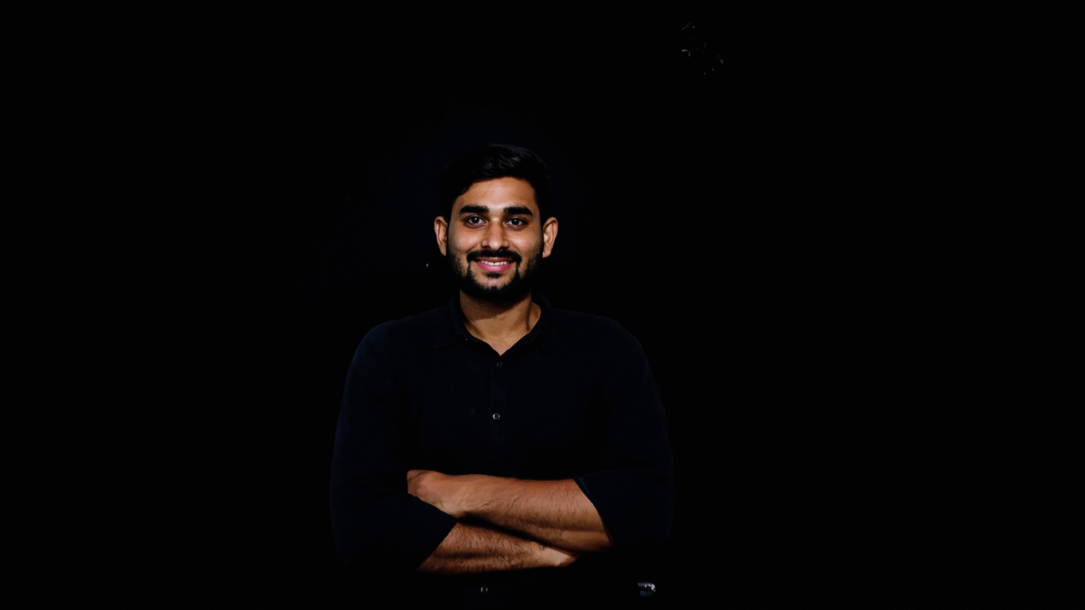 A man with short black hair and a beard, smiling confidently with arms crossed, wearing a black shirt against a dark background